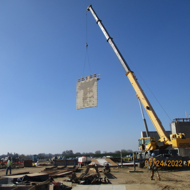 A construction site with a crane lifting a large panel against a clear blue sky.