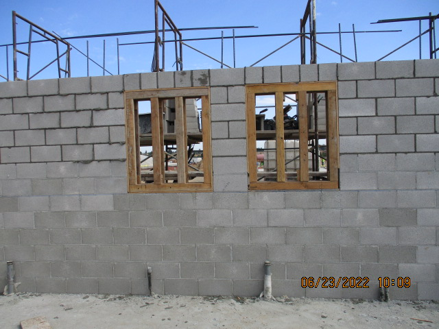 A construction site with a cinder block wall and two wooden window frames, scaffolding visible in the background.