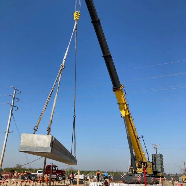 A crane lifts a large concrete beam on a construction site under a clear sky. Workers and vehicles are visible.