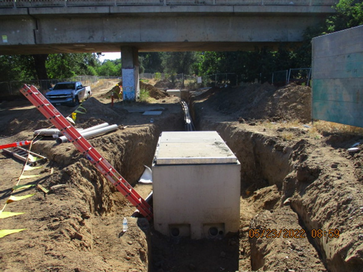 Construction site under a bridge with trenches, conduit pipes, and a ladder.