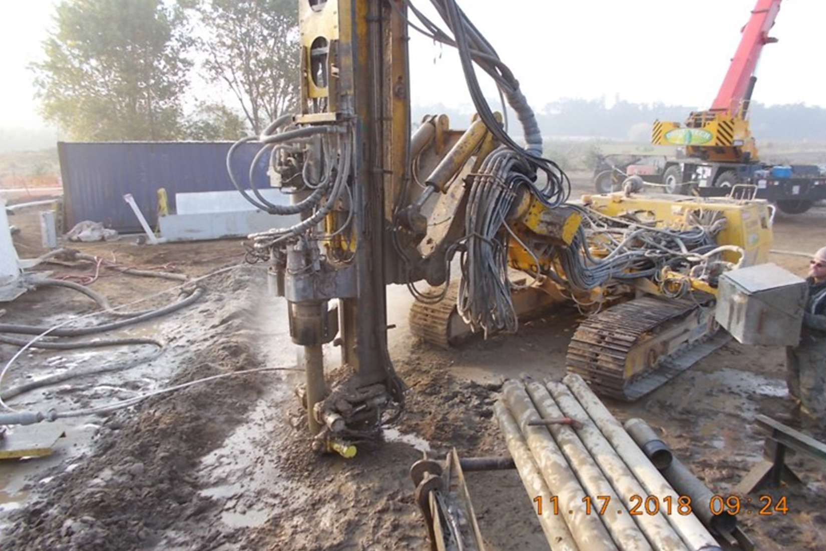 A construction site with machinery, including a drilling rig and a crane, with workers and equipment visible.
