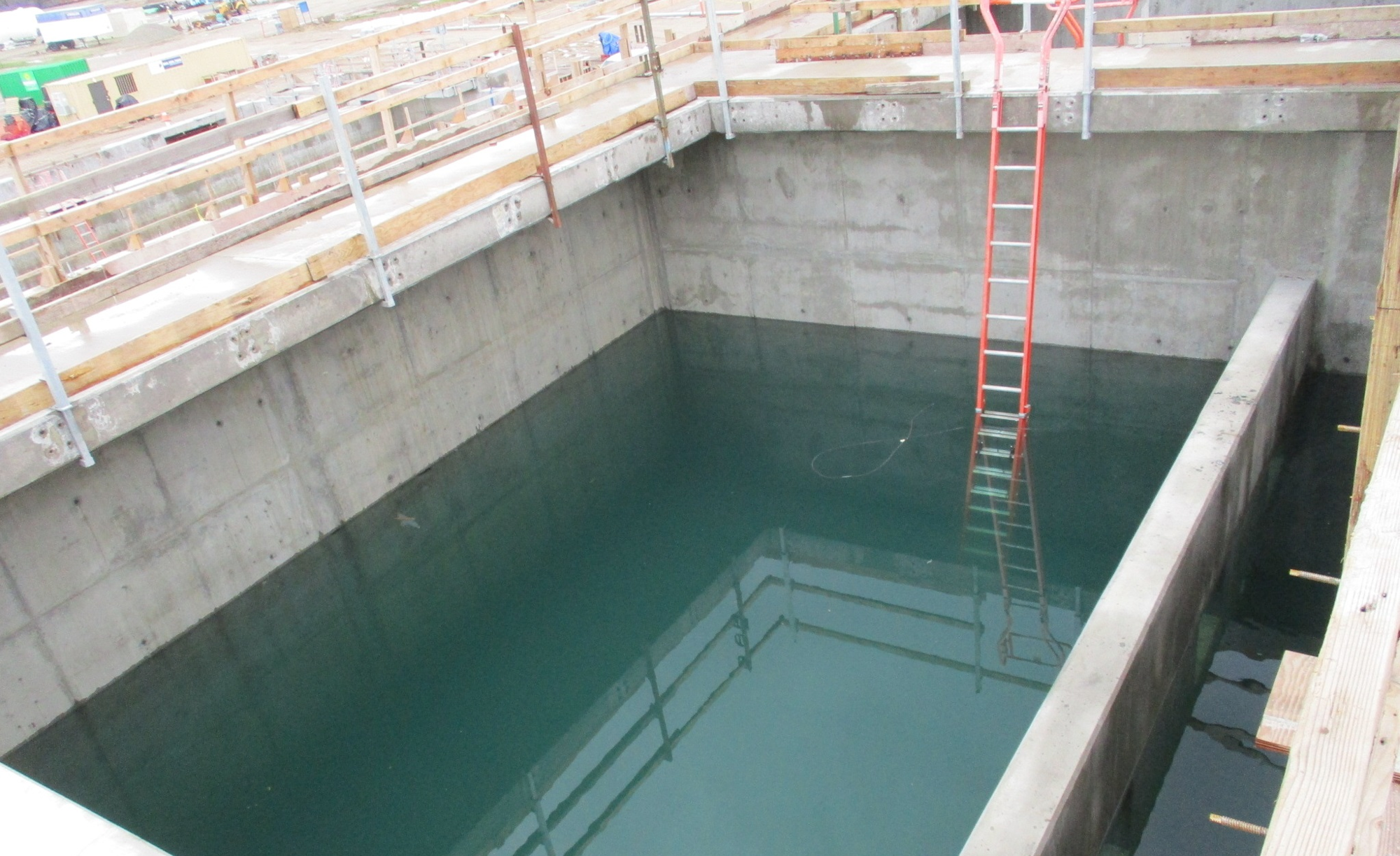 A construction site with a deep, water-filled concrete pit and a red ladder descending into it.