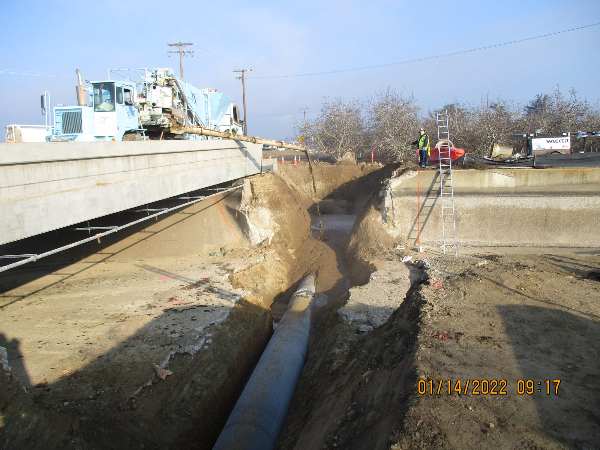 Construction site with a trench, large pipe, machinery, and a worker by a ladder.