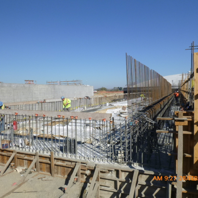 Construction site with workers, concrete structures, steel reinforcement, and scaffolding under a clear sky.
