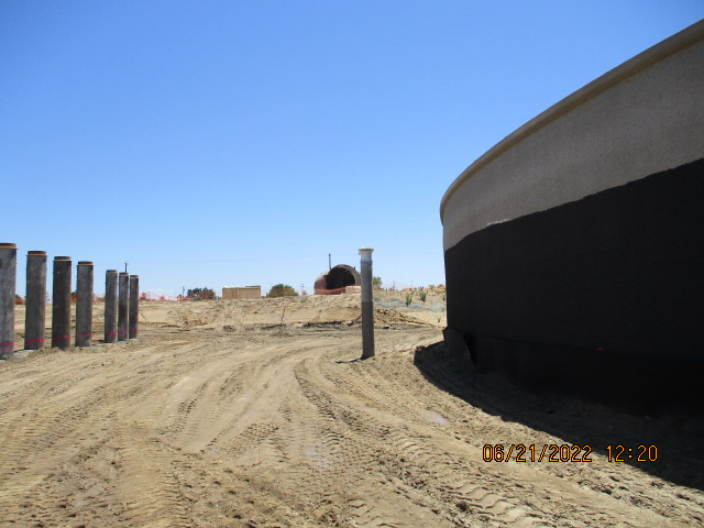 Construction site with tire tracks, a large tank, and vertical pipes in a sandy area under clear skies.