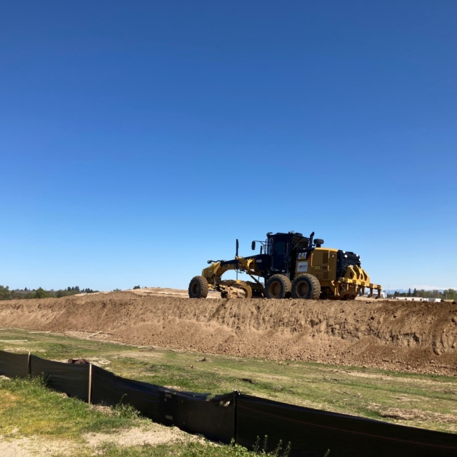 A motor grader working on a dirt surface under a clear blue sky.