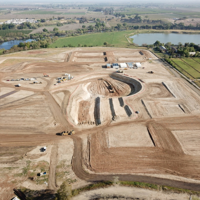 Aerial view of construction site with earthworks and machinery near fields and a body of water.