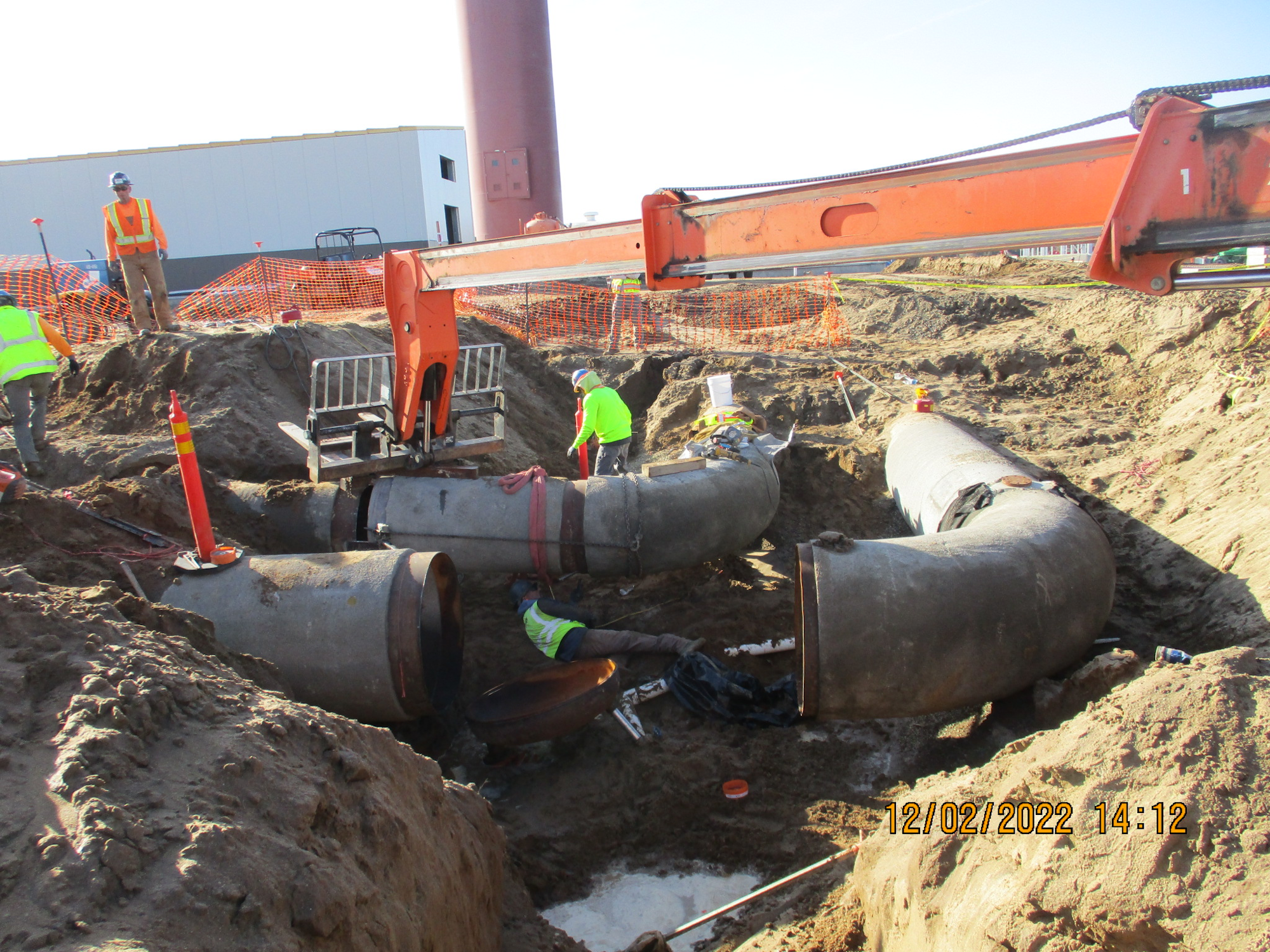 Construction workers installing large pipes in a trench with machinery and safety equipment visible.