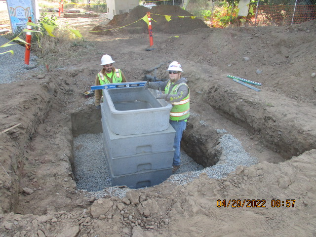 Two workers in high-visibility vests installing an underground utility box at a construction site.