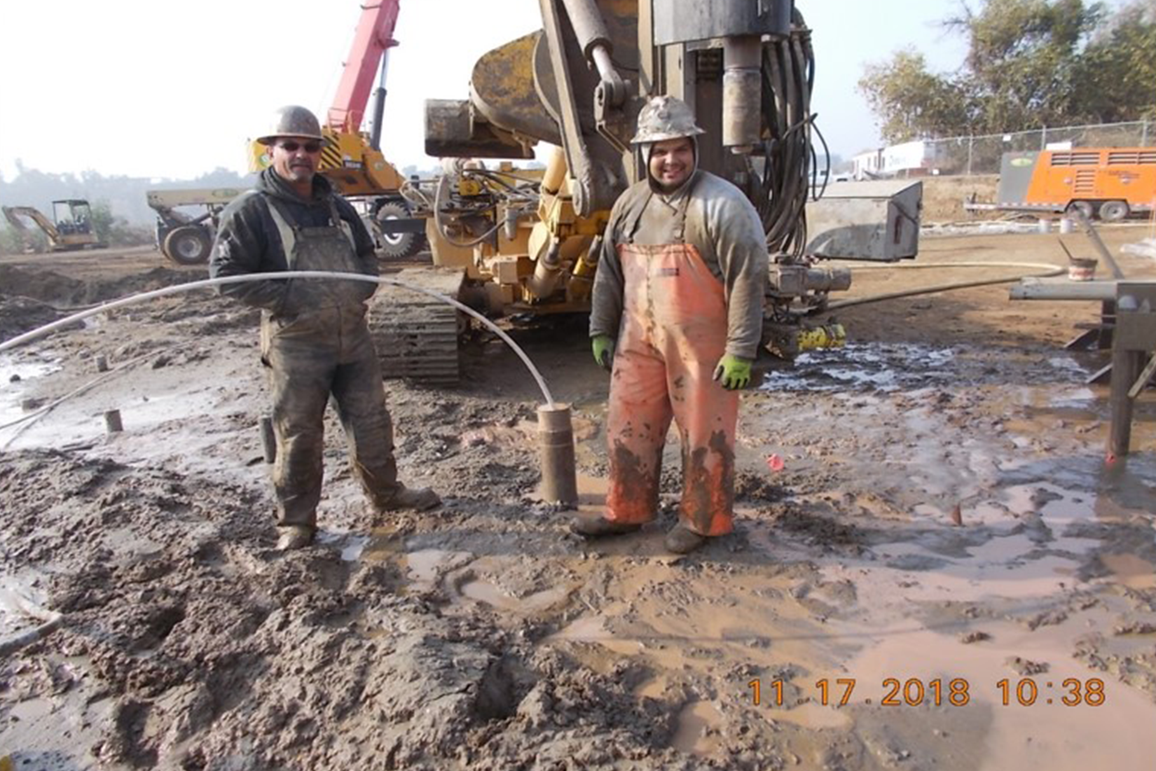 Two workers at a muddy construction site with drilling equipment.