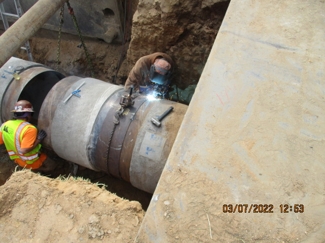 Workers welding a large pipe in an excavation site.