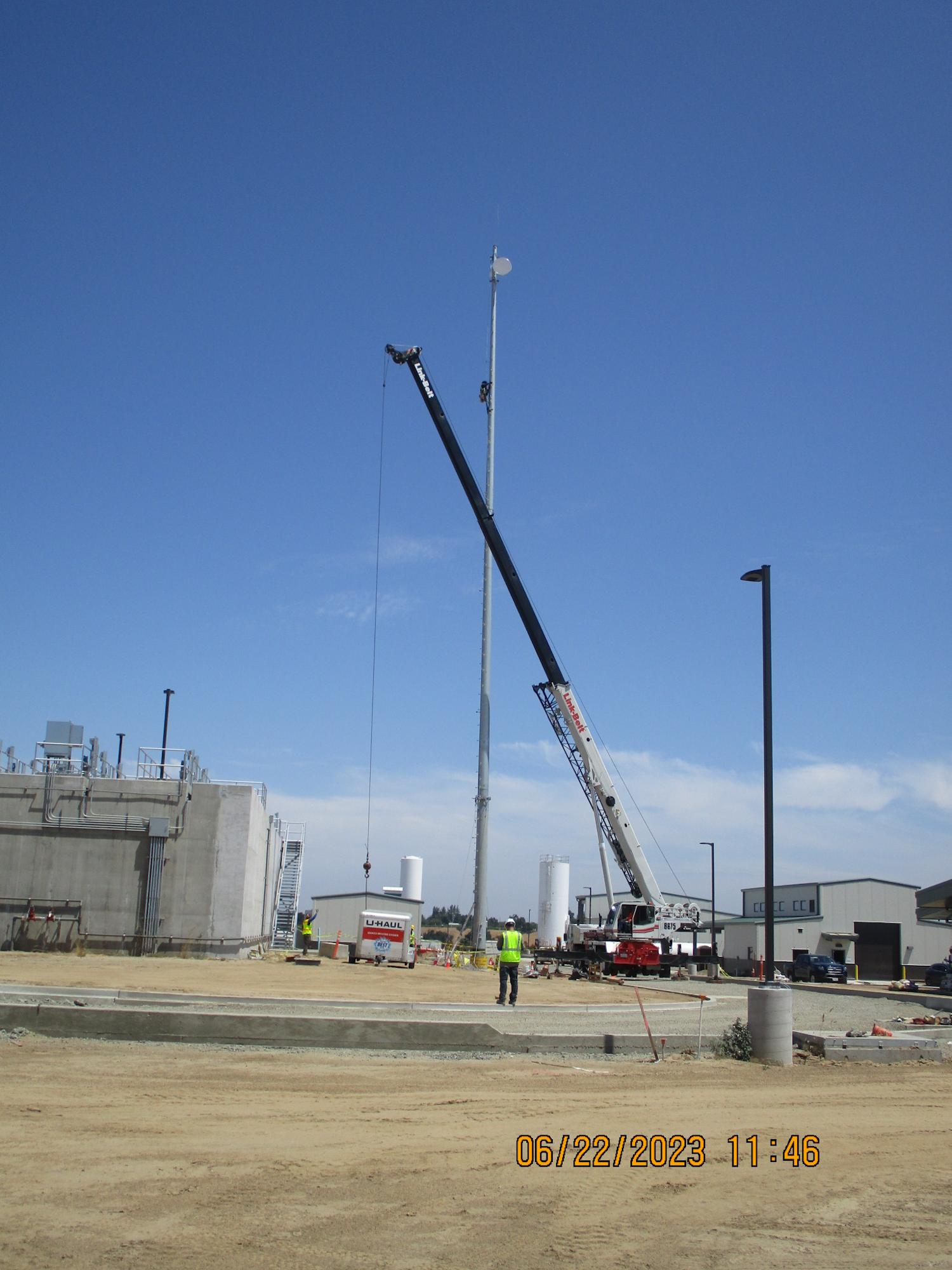 Construction site with a crane lifting an object, workers in vests, clear sky, and industrial buildings.