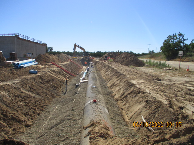 A construction site with a trench for pipeline installation, machinery, and building materials under a clear sky.