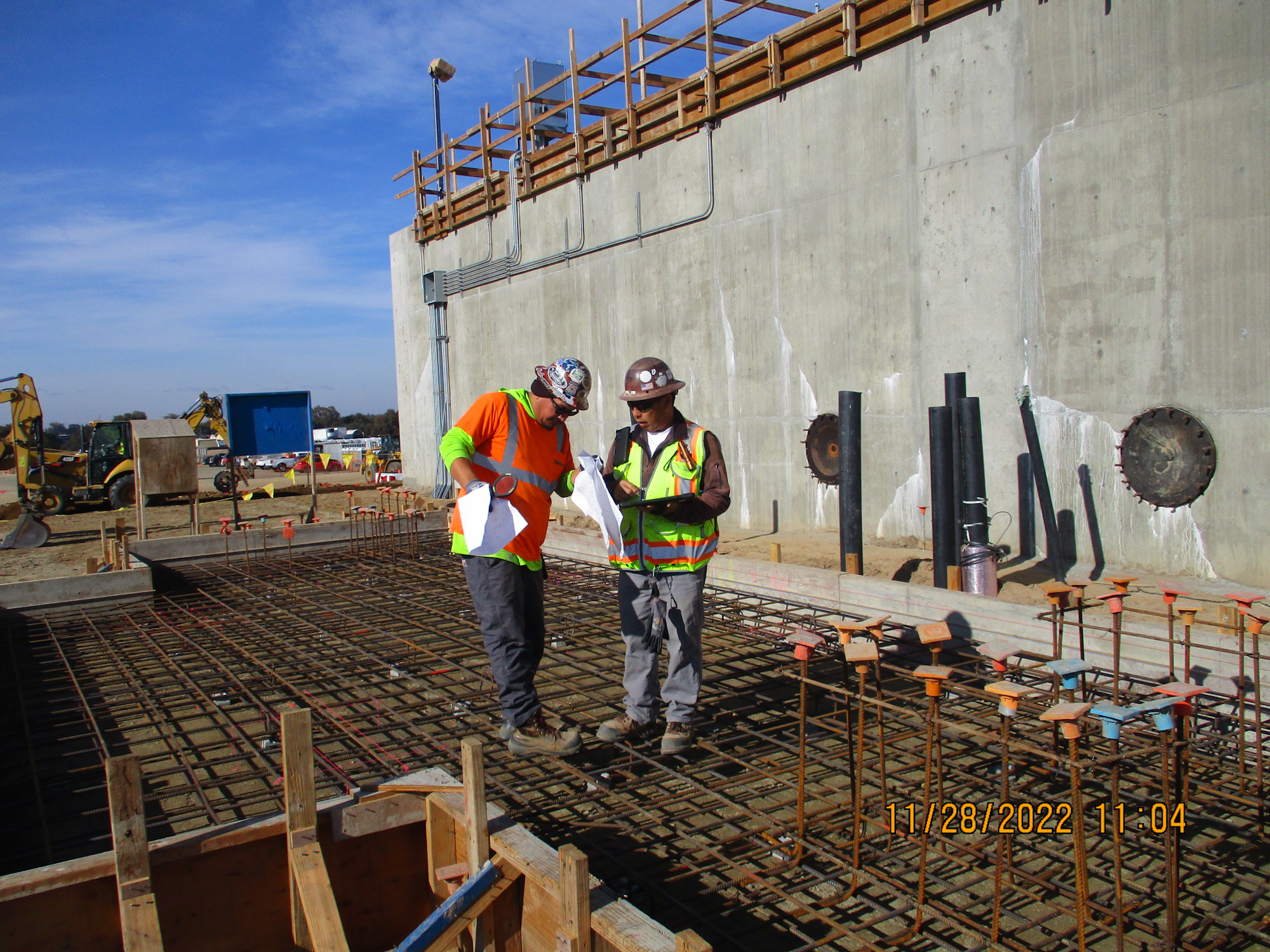 Two construction workers with plans on a rebar-lined foundation, concrete wall, and construction equipment in the background.