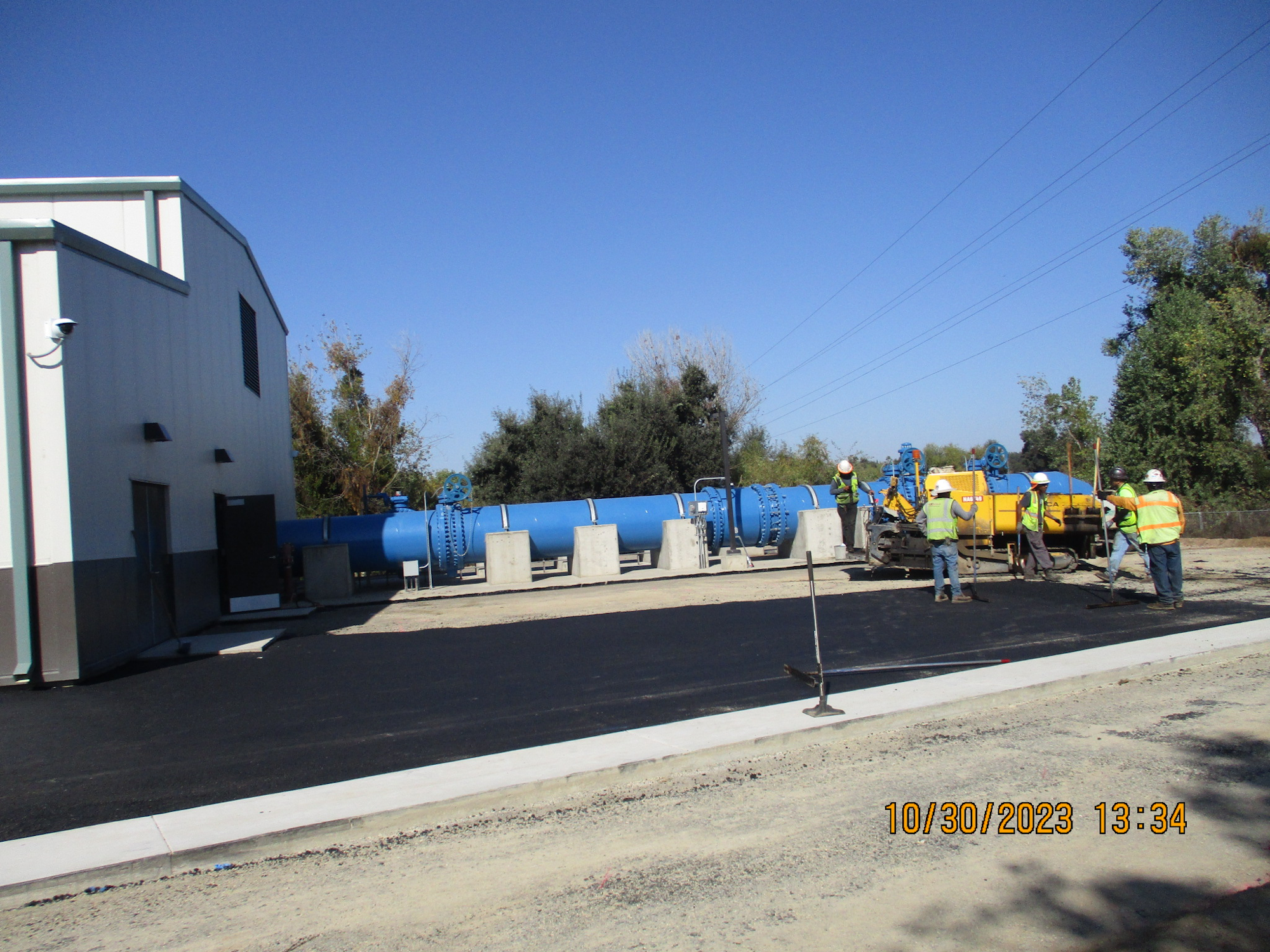 Workers in hi-vis vests at a construction site near a building with large blue pipes and machinery.