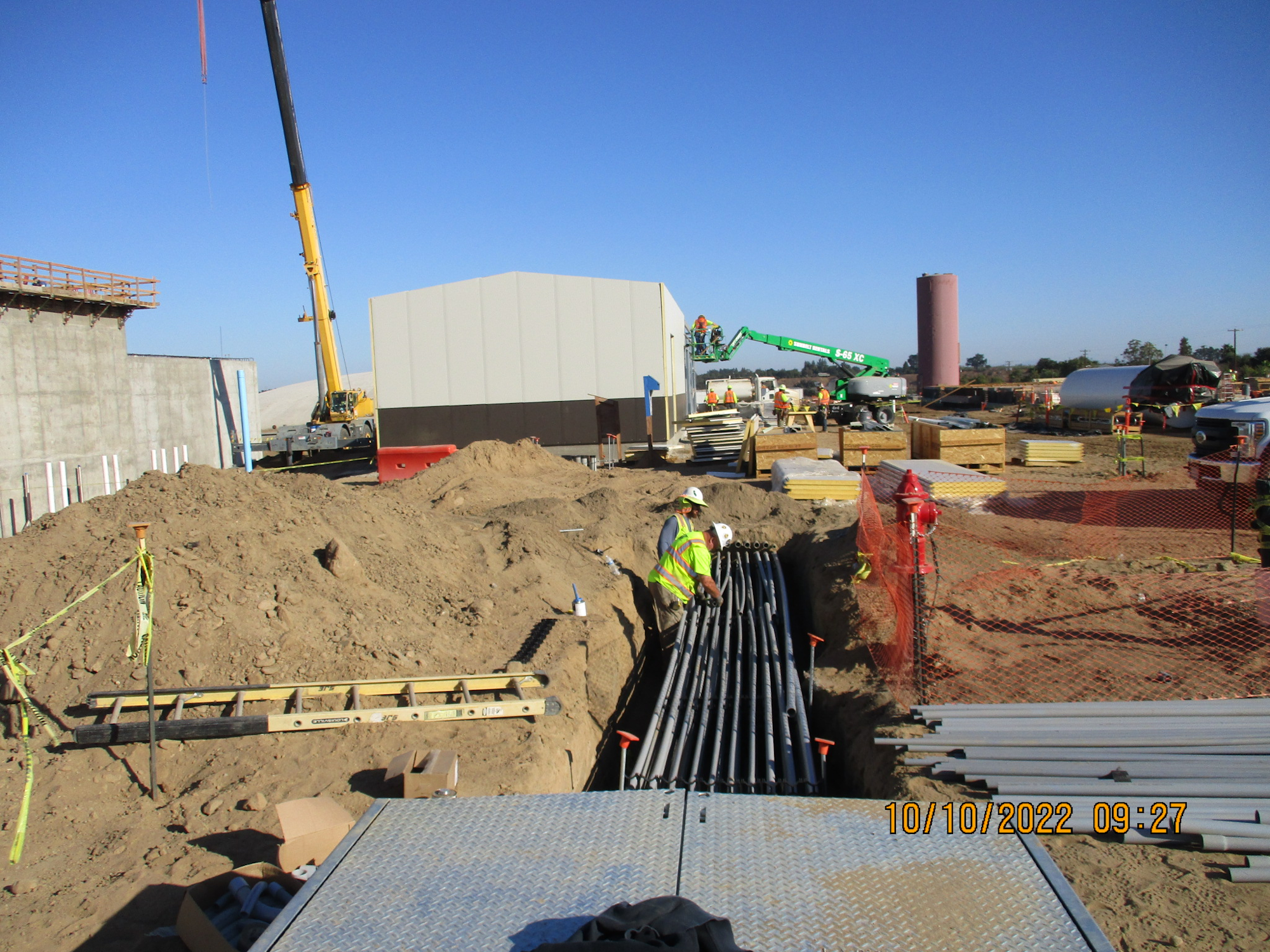 Construction site with equipment, workers, piping, and buildings under a clear sky.