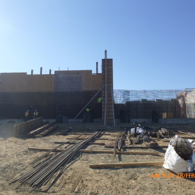 Construction site with workers, concrete forms, rebar, and building materials under a clear sky.