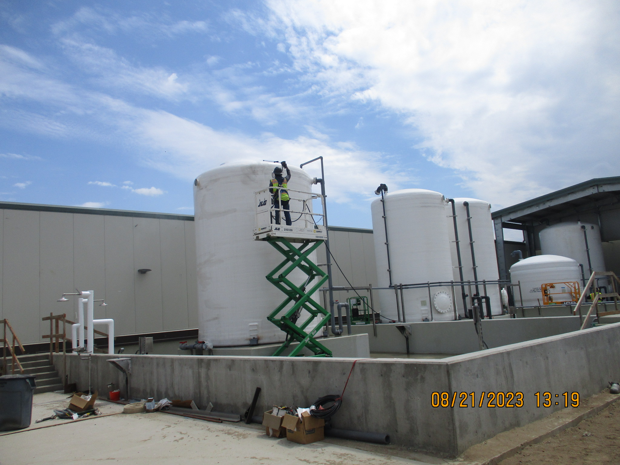 Industrial storage tanks with a person on a lift platform, clear sky, and construction materials on the ground.