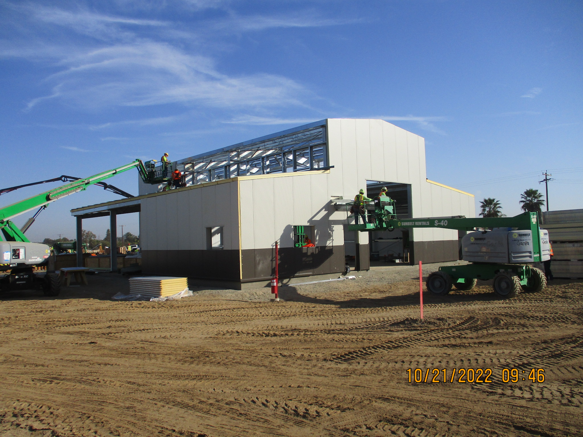 Construction site with workers on boom lifts assembling a building's steel frame under a clear sky.