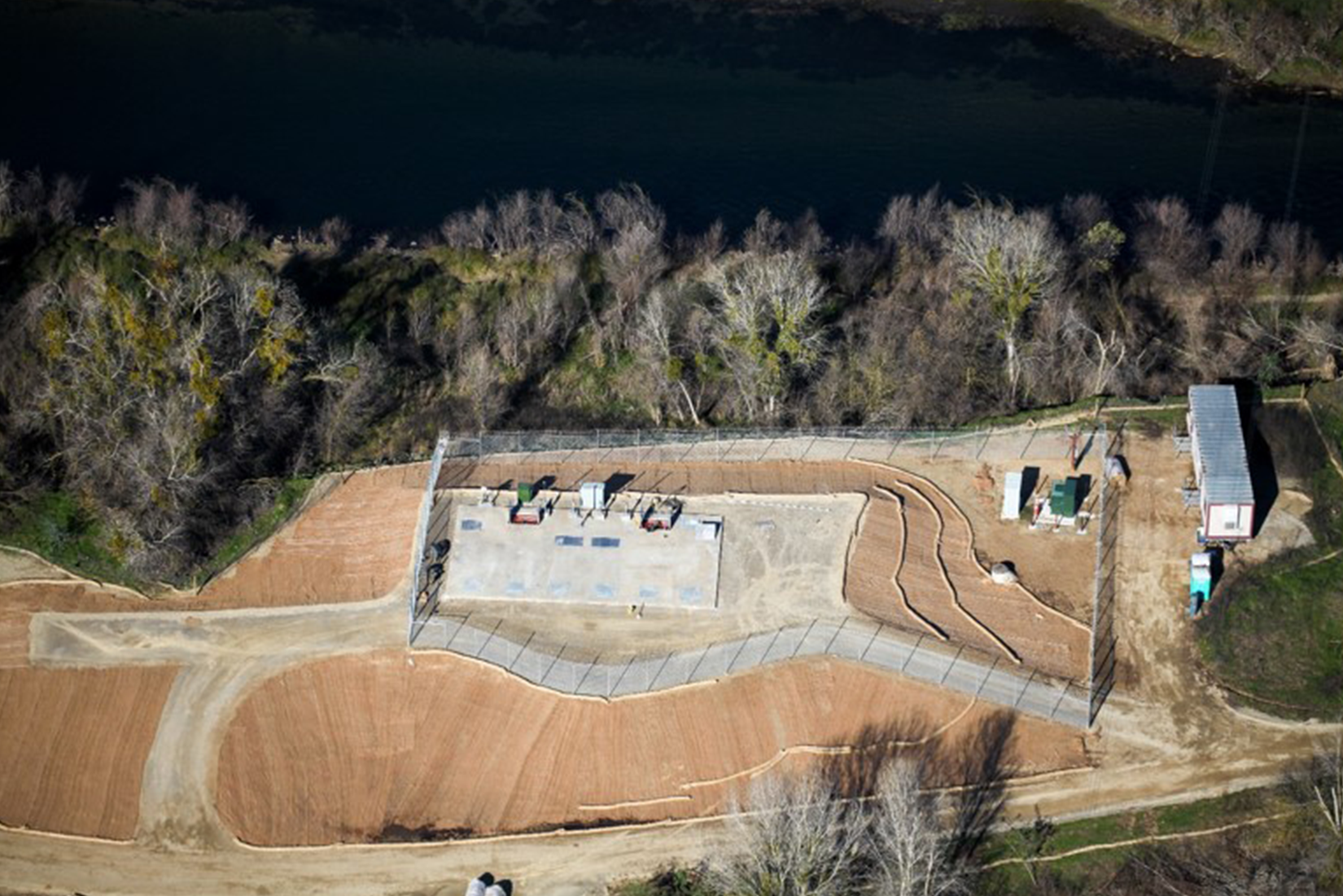 Aerial view of a concrete skateboard park next to a river, with surrounding trees and paths.