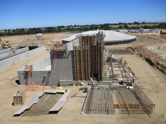 A construction site with workers, scaffolding, excavated earth, and large concrete structures under a clear sky.