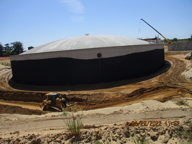 A large circular storage tank at a construction site with a bulldozer and crane visible.