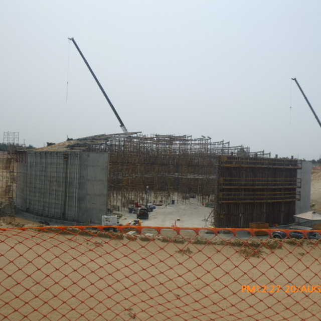 Construction site with cranes, scaffolding, and concrete structures, viewed through an orange safety net.