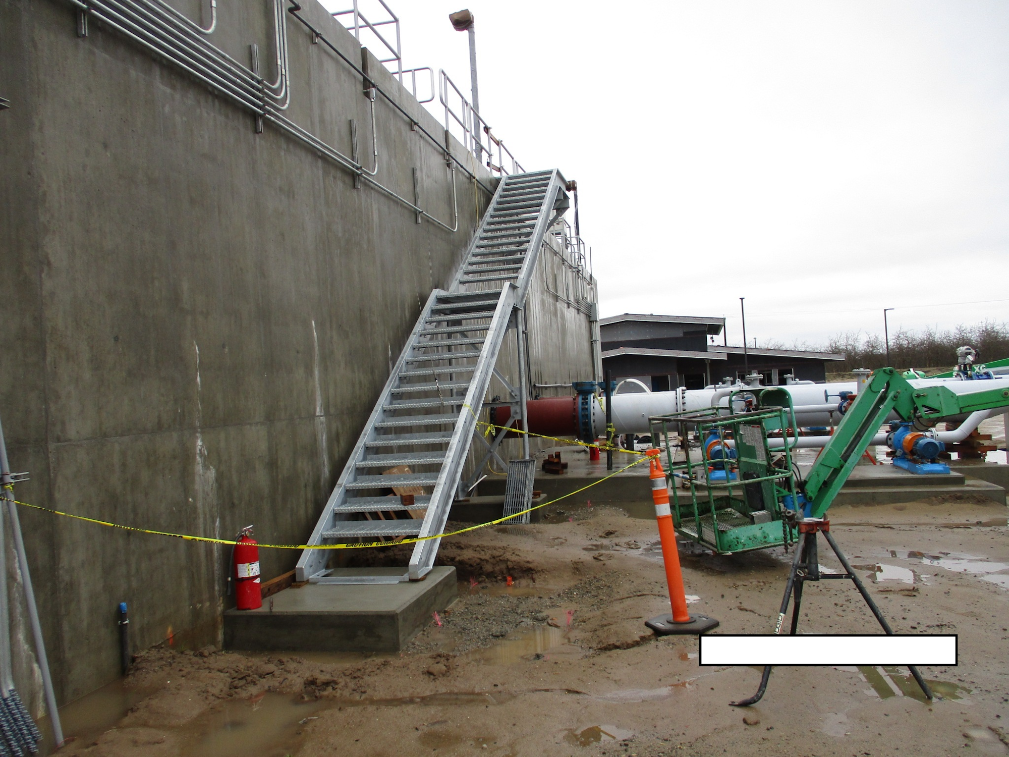 Metal staircase beside a concrete wall, industrial equipment, caution tape, and muddy ground.