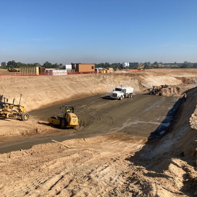 A construction site with heavy machinery and vehicles, excavated land, and buildings in the background.