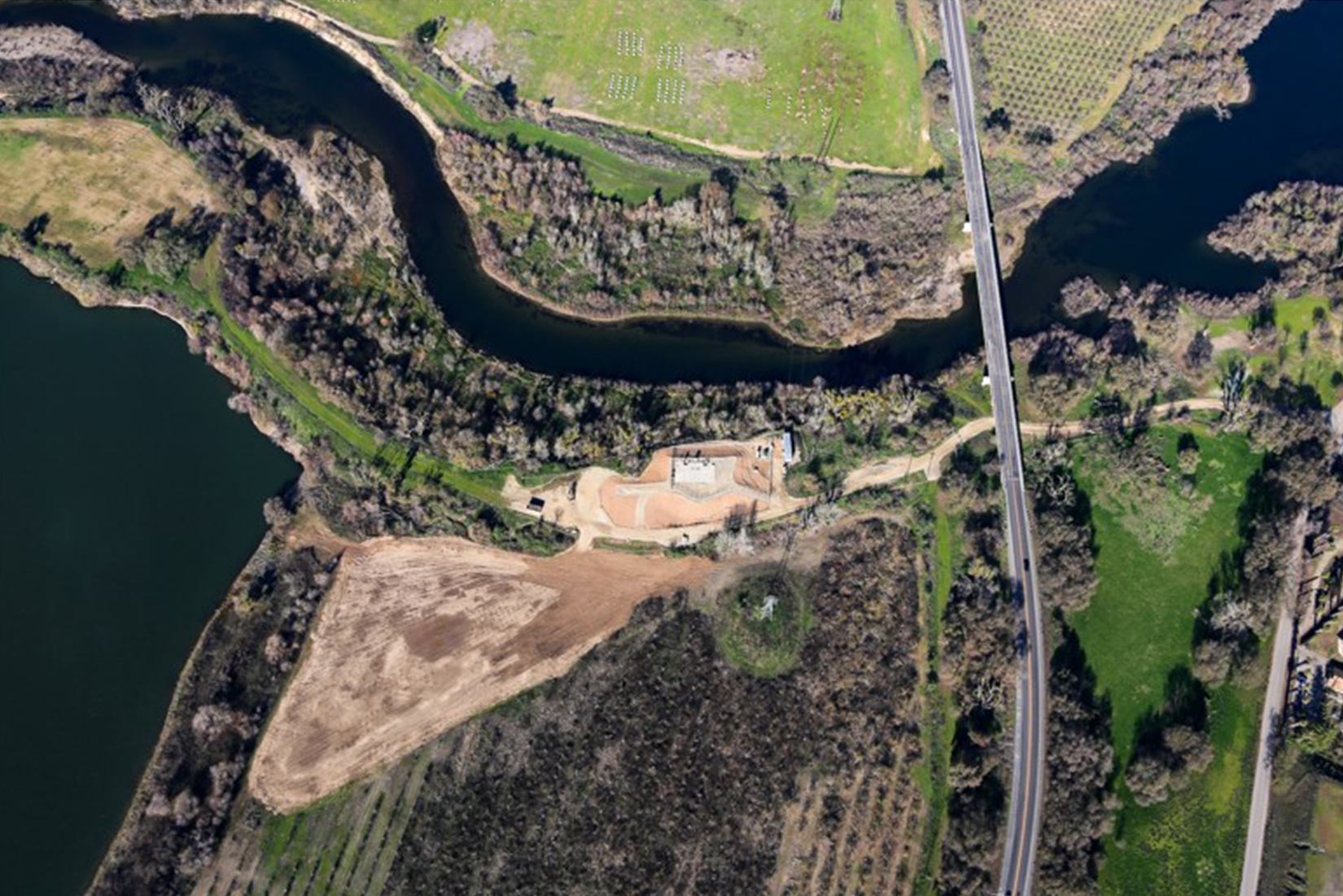 Aerial view of a meandering river with adjacent roads, fields, and a small building complex.