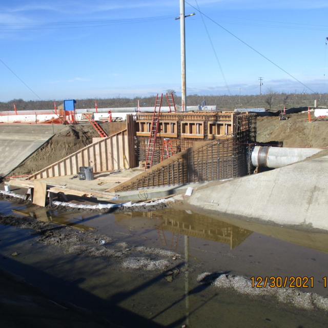 A construction site with concrete structures, rebar, wooden forms, standing water, and a ladder.