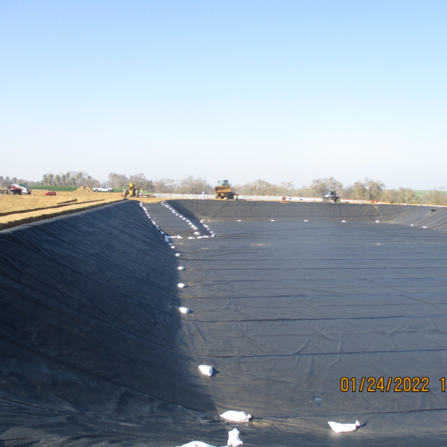 Large pond or pit lined with black geotextile, sandbags placed in a row, machinery and people in the background.