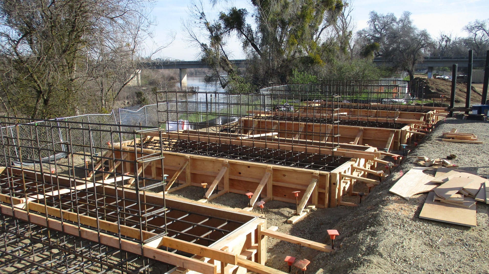Construction site with wooden formwork and steel rebar for concrete foundation near a river.