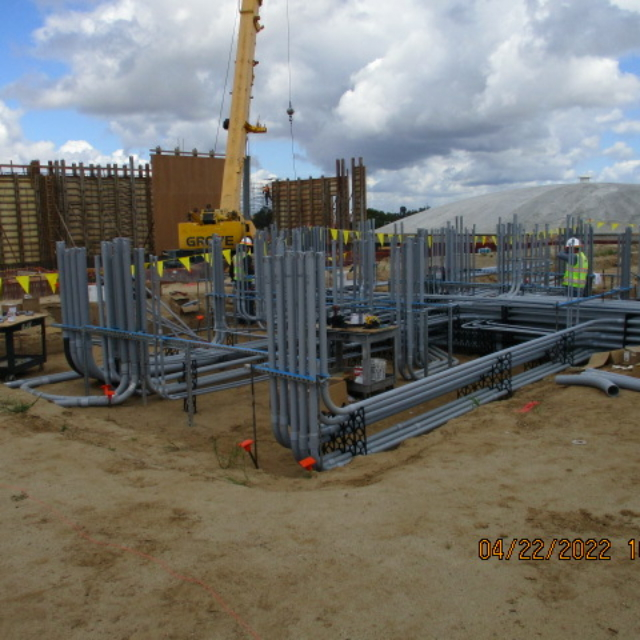Construction site with steel structures, conduit piles, workers, and crane under cloudy sky.
