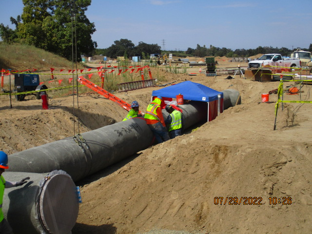 Construction site with workers installing a large pipe underground, safety barriers around, vehicles in the background, and a timestamp.