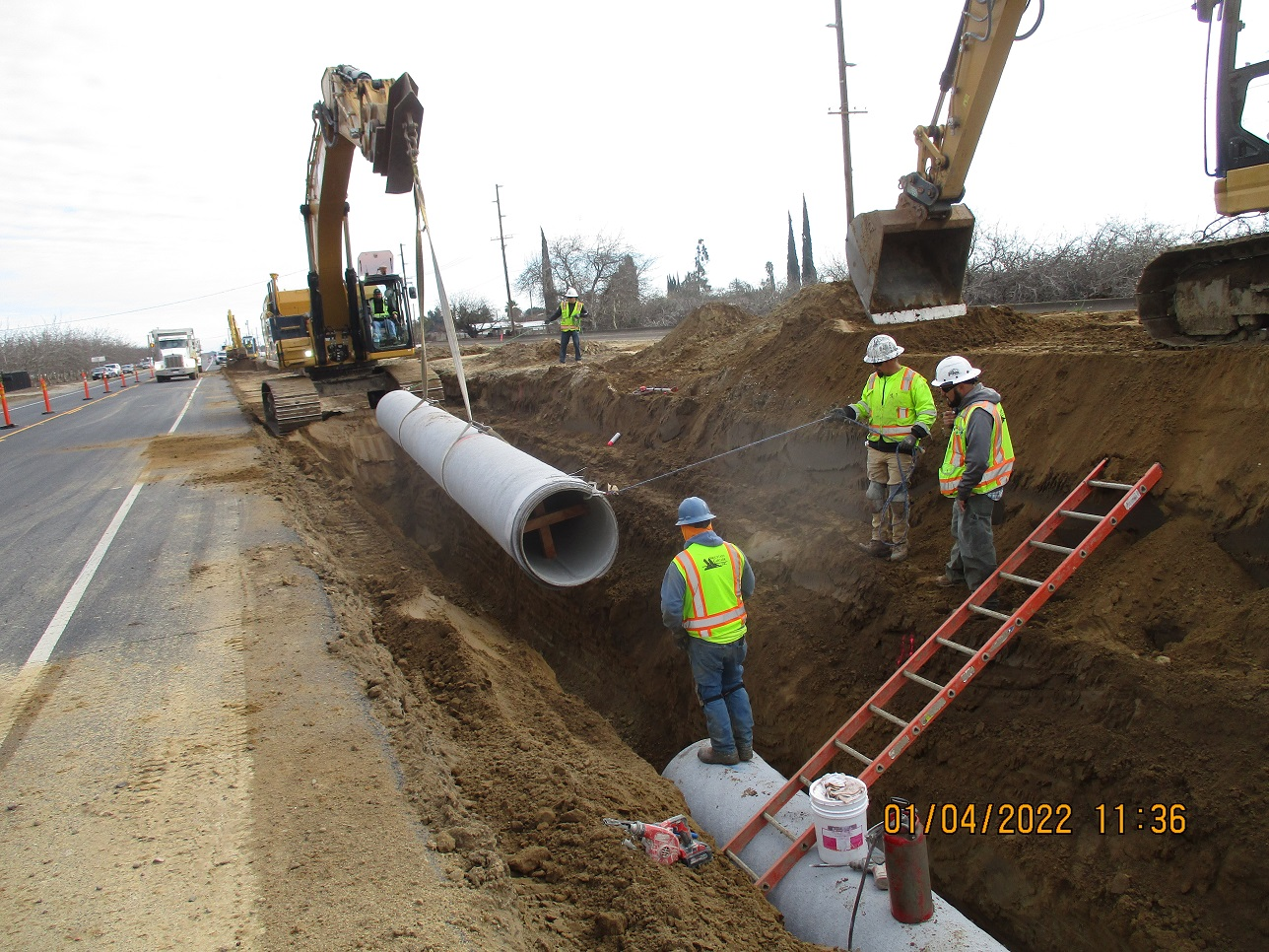 Construction workers installing a large pipe in a trench with an excavator nearby.