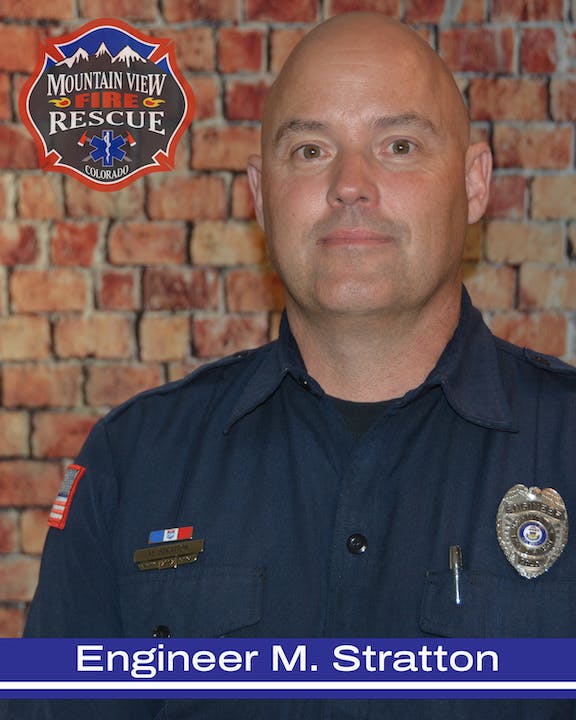 A firefighter in uniform with badges, in front of a brick wall with a logo for Mountain View Fire Rescue, Colorado.