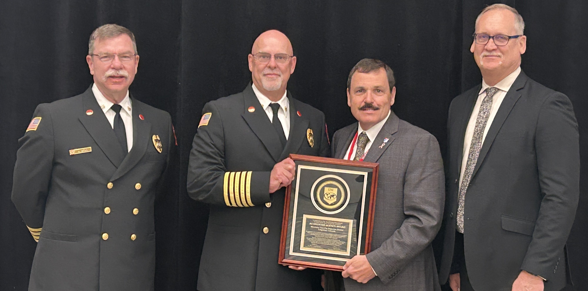 Four men in formal attire, two in fire chief uniforms, holding an accreditation award plaque.