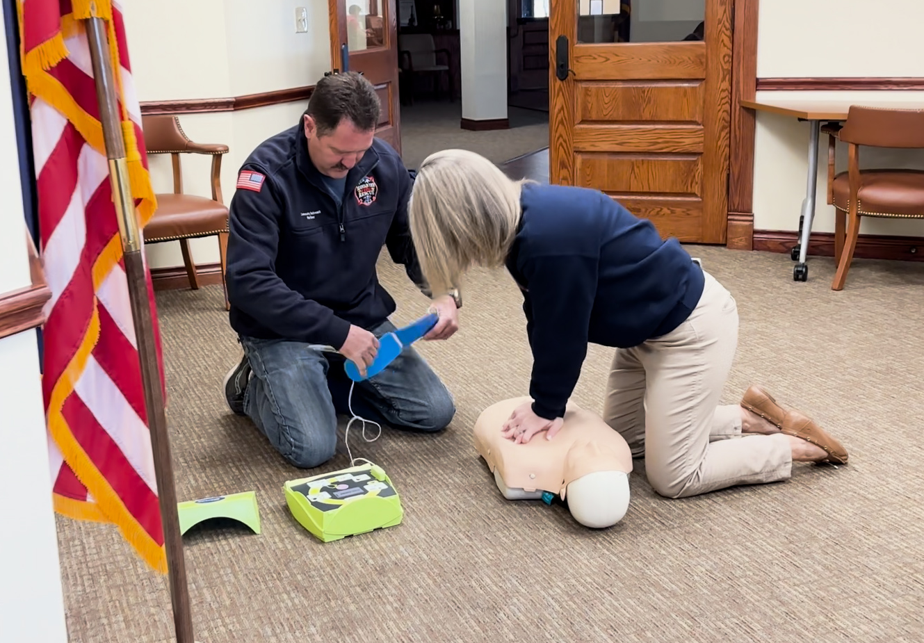 A man and woman are practicing CPR and using a defibrillator on a mannequin in a room.