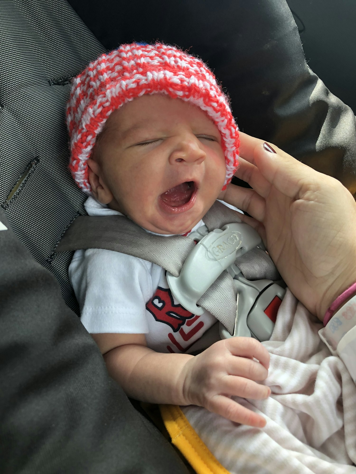 A newborn baby in a car seat yawning, wearing a knitted hat, while being held by an adult's hand.