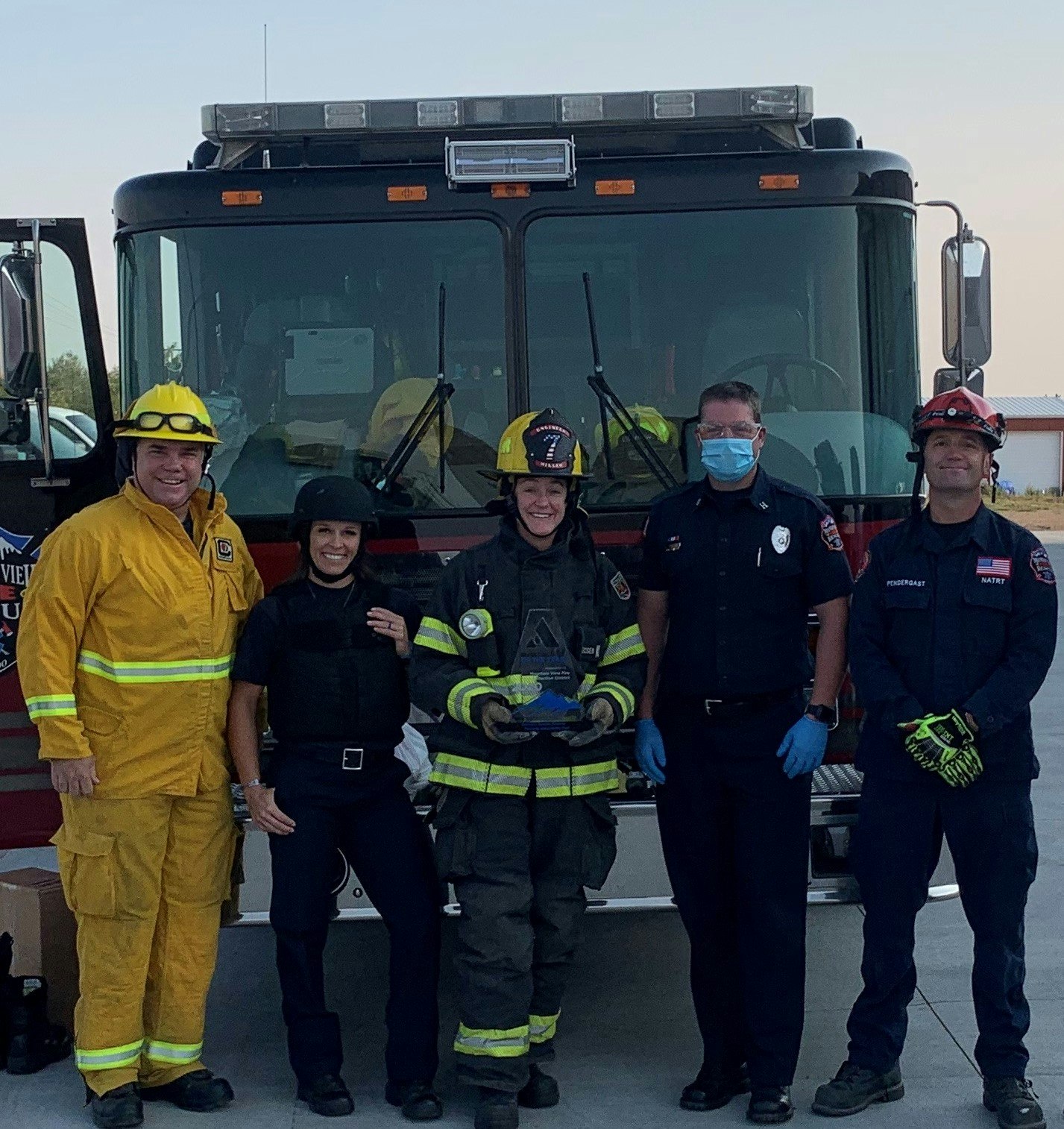 A group of firefighters and officials stand in front of a fire truck, celebrating an award with smiles and safety gear.