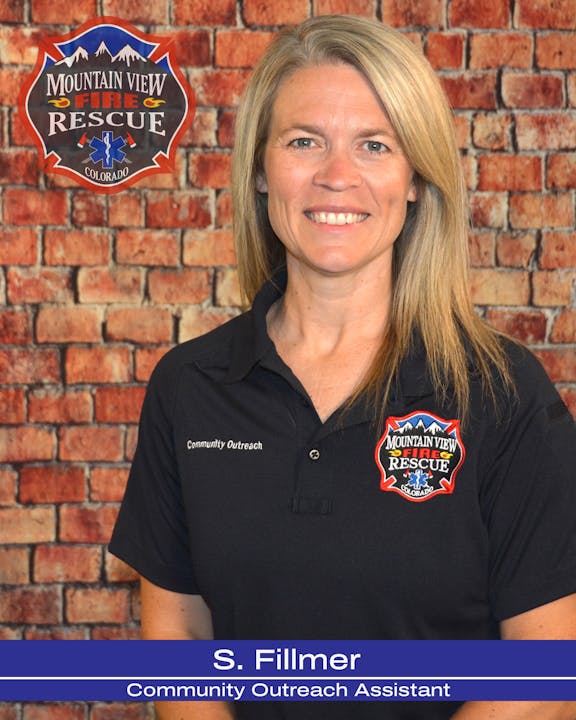 A smiling woman in a black shirt with a "Community Outreach" badge, standing in front of a brick wall backdrop.
