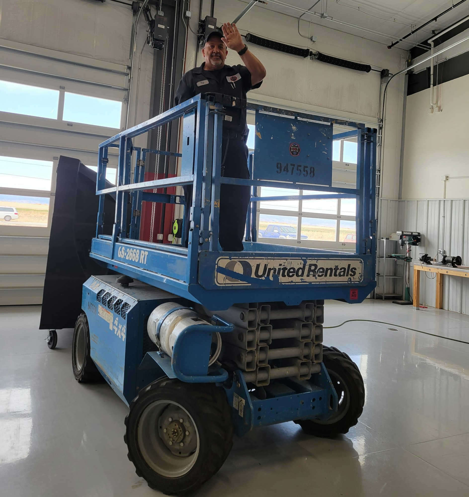 A man waves from a blue scissor lift inside a spacious garage with equipment and large windows.