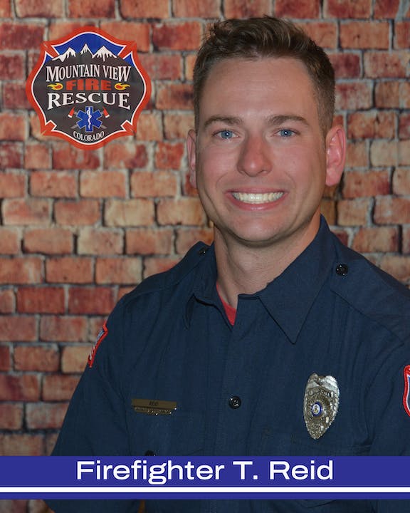 A smiling firefighter in uniform with a badge and nameplate, in front of a brick background with a fire department logo.