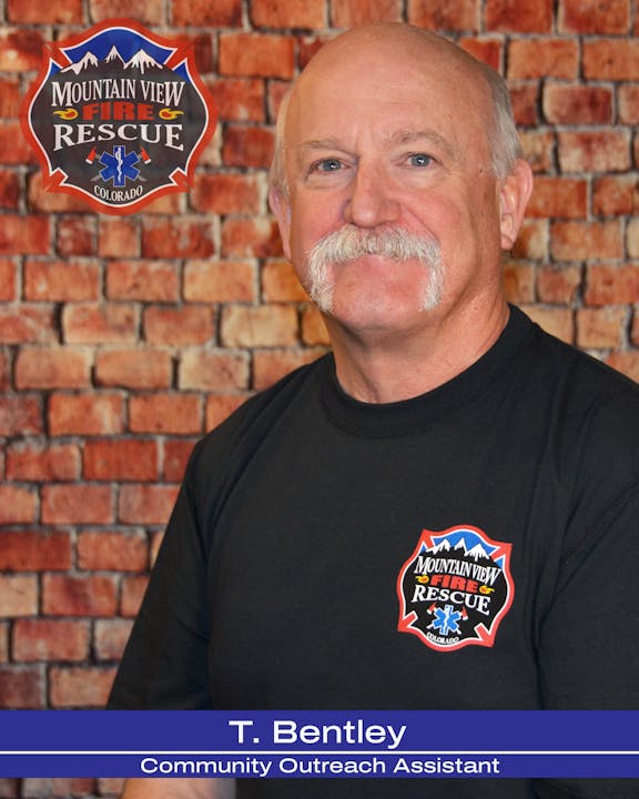 A man with a mustache, wearing a Mountain View Fire Rescue shirt, stands in front of a brick wall.