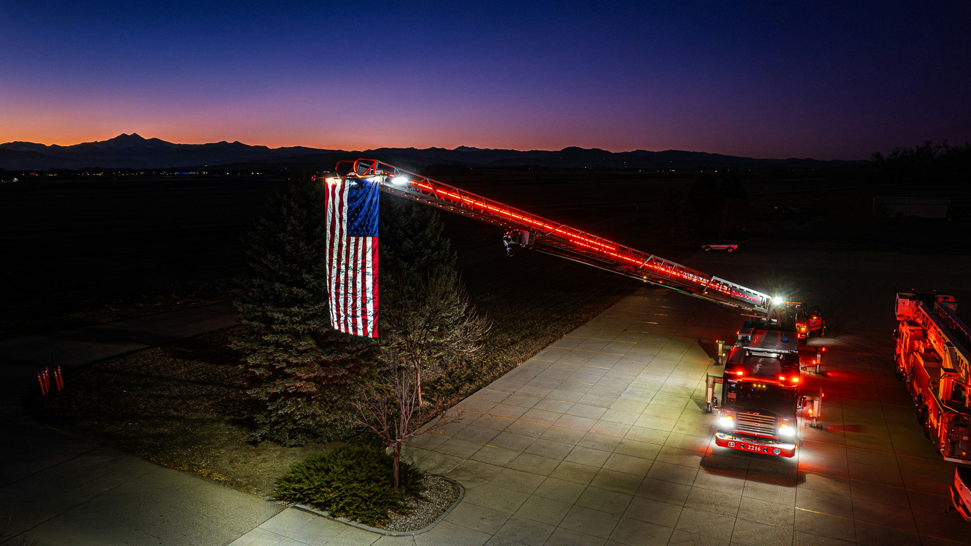 Fire truck with an extended ladder displaying a large American flag at dusk.