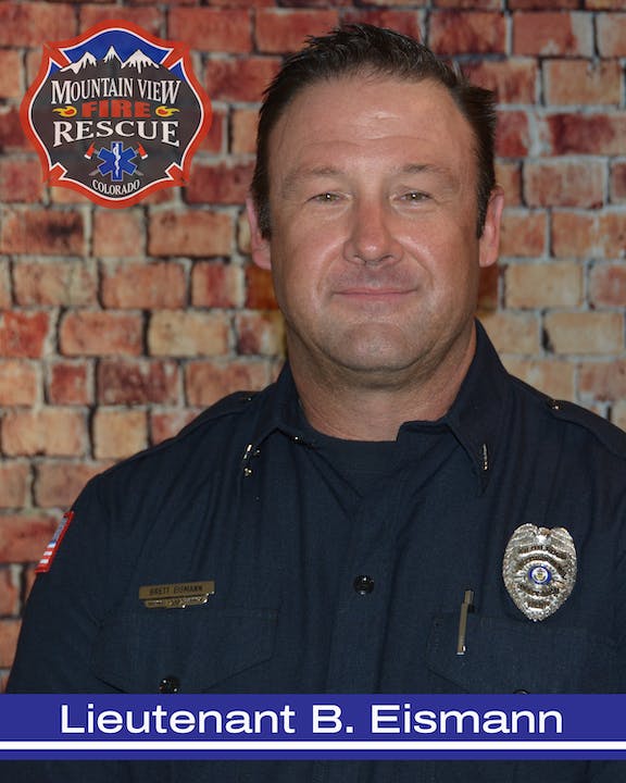 A firefighter in uniform with an emblem and a nametag, standing against a brick wall background.