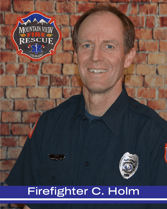 Firefighter in uniform with badge, smiling in front of a brick wall, with a fire and rescue emblem above.