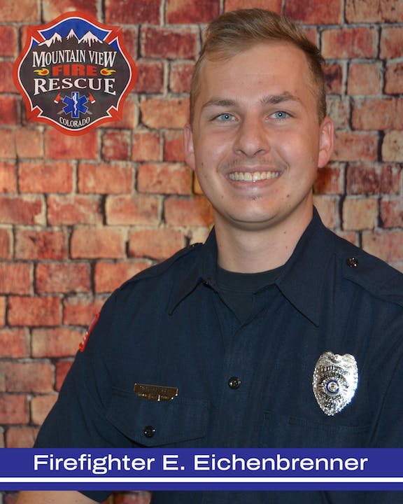 A smiling firefighter in uniform with a badge, brick background, and fire rescue emblem.
