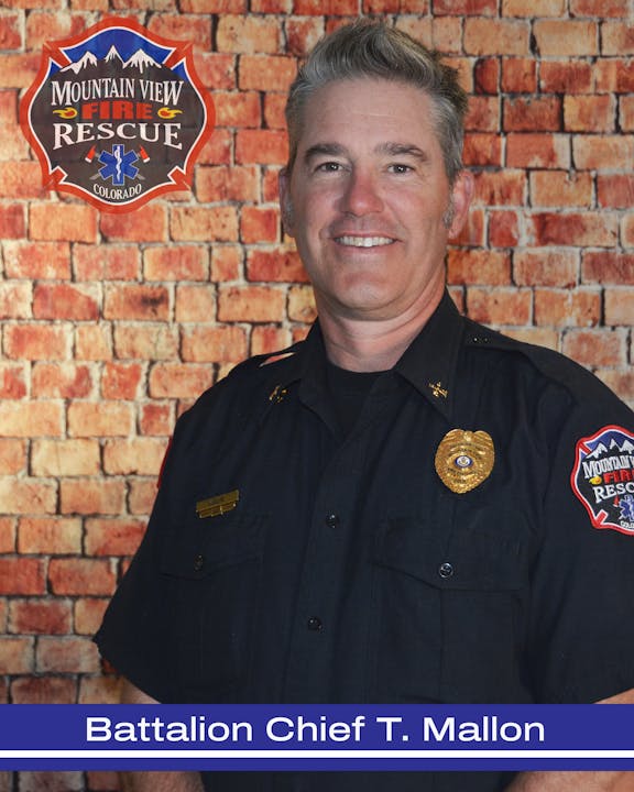 A smiling firefighter in uniform with a badge, in front of a brick wall. Text: "Battalion Chief T. Mallon, Mountain View Fire Rescue Colorado."